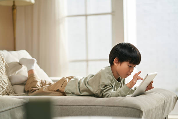 little asian boy lying on front on couch playing computer game using digital tablet stock photo