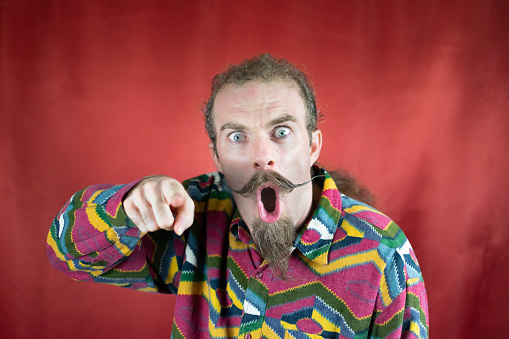 Angry hipster man pointing at camera amazed and with surprised expression with red background