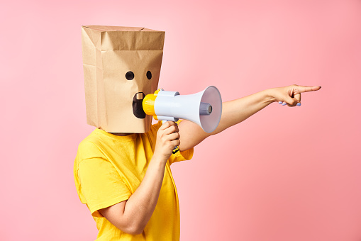 Anonymous female with paper bag on head screaming in megaphone and pointing finger to the side over pink background