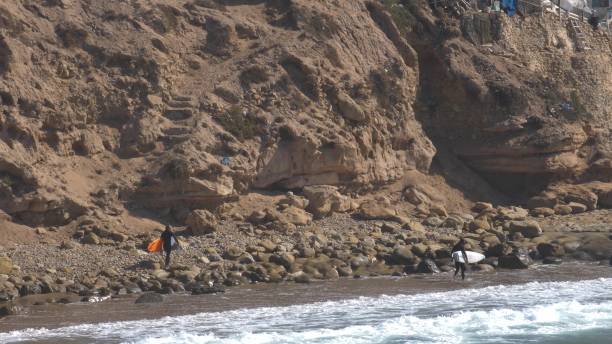 istock Surfers walking on Atlantic Ocean Coast with Cliffs and Small Village in Morocco 1644320984