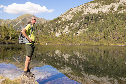 A gray-haired man with a backpack watches a mountain lake with a perfect reflection, Austria. High quality photo