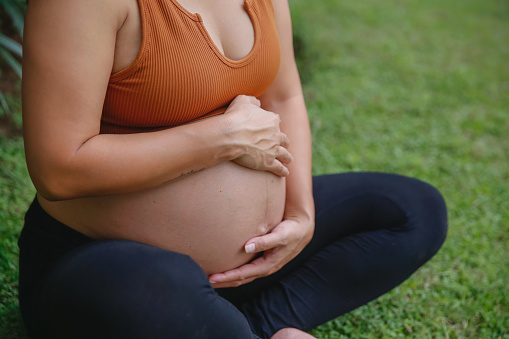 Close-up front view shot of unrecognizable pregnant Indonesian woman feeling pregnancy contractions while sitting on the grass crossed legged. There's a linea nigra dark line on her stomach.