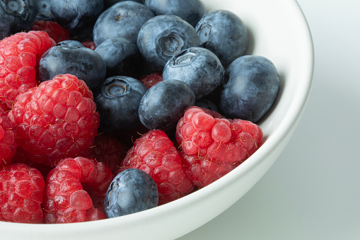 Closeup view of fresh raspberries and blueberries in a white bowl on a white background