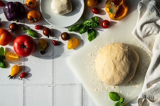 Step by step process of vegetable focaccia baking. Ball of homemade dough and fresh colorful tomatoes, peppers, basil and mozzarella on white tile background.