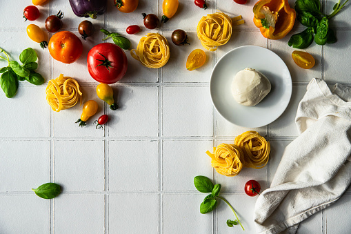 Dry tagliatelle nests, variety of fresh tomatoes, peppers, basil and mozzarella ball on plate on white tile background.
