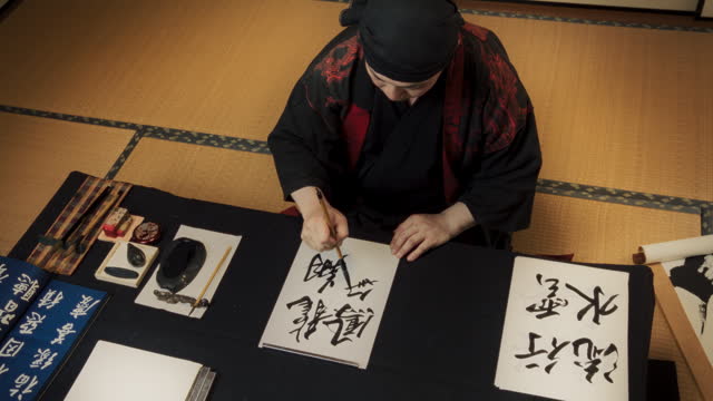 High Angle Footage of a Japanese Artist Working on a Commissioned Calligraphy Work for a Client. Adult Calligrapher Sitting in a Traditional Studio, Writing Kanji Symbols with Ink on a Piece of Paper