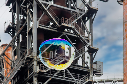 Low angle view of a colorful spherical shaped large soap bubble floating in front of an abandoned and dilapidated tower of an industrial steel mill in Landschaftspark, Duisburg, Germany