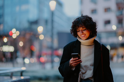 Wide shot of a joyful dark haired young woman standing on the street in a city center and looking down to her smart phone. Having a big smile.Blurred background of a street and city lights.Copy space.