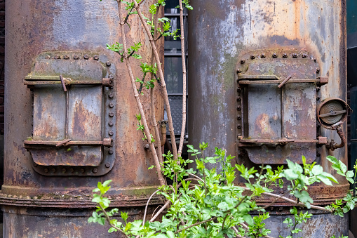 Close up of rusted and weathered furnace or piping system with steel doors of an abandoned and dilapidated industrial steel mill in public Landschaftspark, Duisburg with some green growing in front