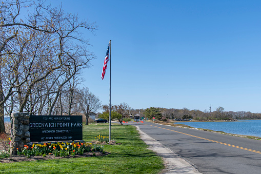 Old Greenwich, CT, USA-May 2022: View of the entrance of Greenwich Point Park or Tods Point with large entrance sign next to road with American flag against a blue sky