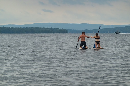 Meredith, United States – July 22, 2023: Two people, standing atop paddle boards, leisurely enjoy the tranquil waters of a serene lake