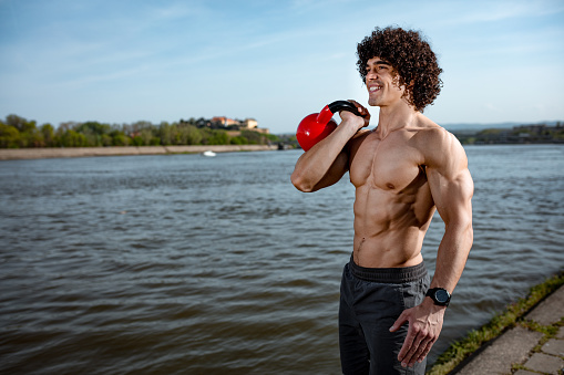 Bodybuilder with six pack, perfect ABS, shoulders, biceps, triceps and chest  is doing strong fit body training with kettlebell, near the river.