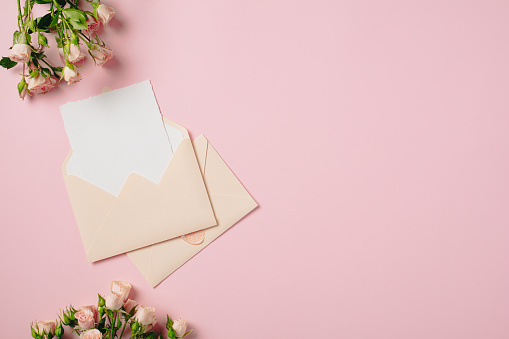 Wedding invitation card inside envelope with roses flowers on pink background. Flat lay, top view, copy space.