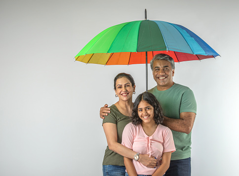 Portrait of smiling mature father holding umbrella and protecting mother and daughter during rainy season over white background