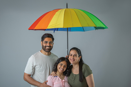 Portrait of smiling mature father holding umbrella and protecting mother and daughter during rainy season over white background