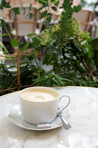 Photo of a cup of latte coffee, chocolate and spoon on a white marble table next to plants on a terrasse of a café in Paris.