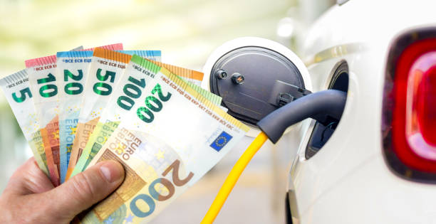 Mano che tiene le banconote in euro davanti a un'automobile elettrica durante la ricarica - foto stock