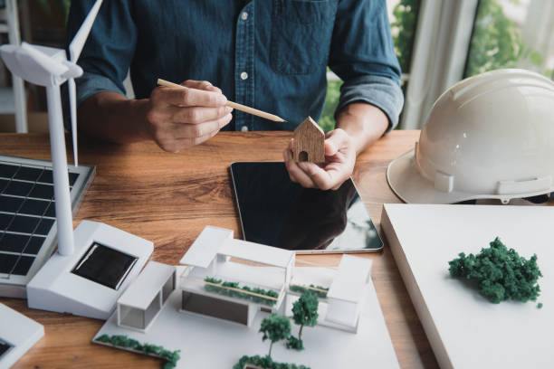 istock Architect designer hold small wooden house symbol while thinking about home design combined with living using natural energy concept, with modern home and wind turbine model, solar panels on desk. 1644487848