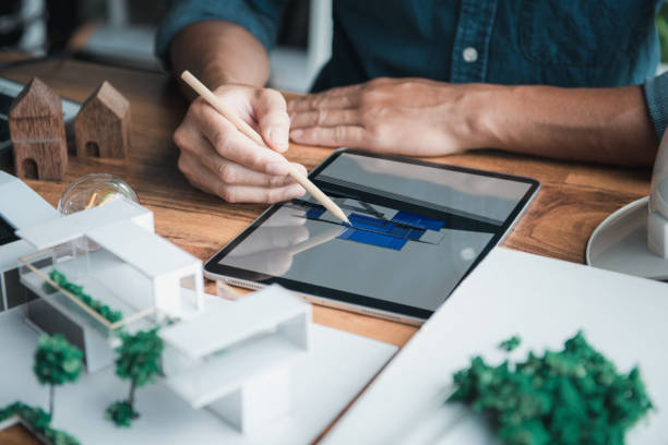 Architect designer holds a pencil pointing to the 3D home office drawing on the tablet while thinking and considering detail in the building, Interior design and landscape around. stock photo