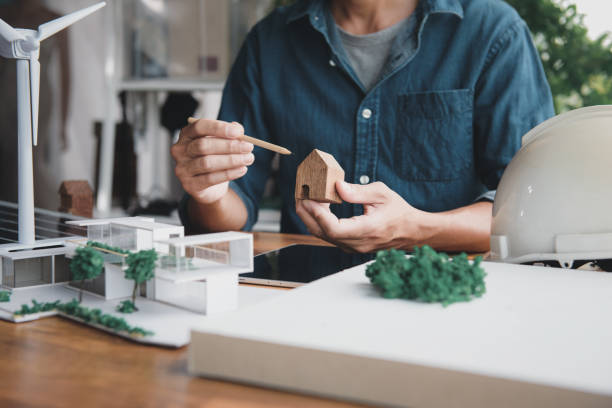 istock Architect designer hold small wooden house symbol while thinking about home design combined with living using natural energy concept, with modern home and wind turbine model, solar panels on desk. 1644487856