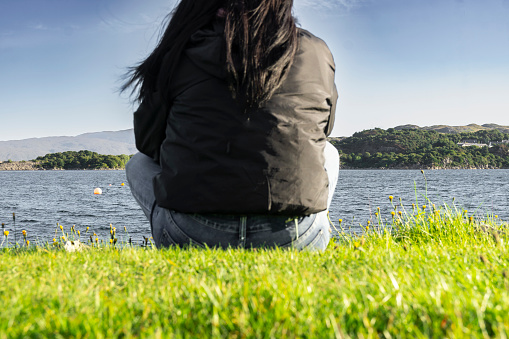 Woman with long brown hair sitting on the grass looking out to sea in Kyleakin Bay, Isle of Skye, Scotland. Vertical shot.