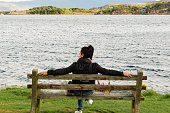 Back shot of a young brown-haired Caucasian woman sitting on a wooden bench, looking relaxed at the sea in Kyleakin Bay on the Isle of Skye. Selective focus on background.