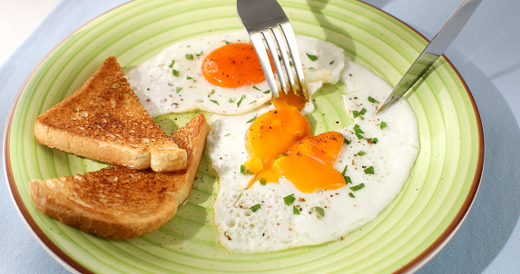 man cuts fried eggs with a knife on a green plate. close-up. angle view. camera tracking. colored background. traditional breakfast with toast . sunny day.