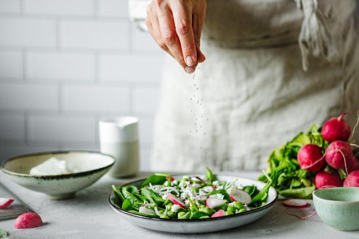 Close-up of a woman hand making healthy summer green salad. Woman prepare healthy green salad using radish, green peas, baby spinach and springing black pepper in kitchen.