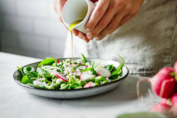 Close-up of a woman preparing healthy summer green salad at home kitchen. Woman adding olive oil on plate of chopped vegetables over kitchen counter.