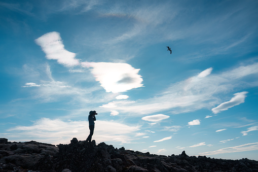 Silhouette of photographer standing with camera taking a photo on the mountain and bird flying in blue sky