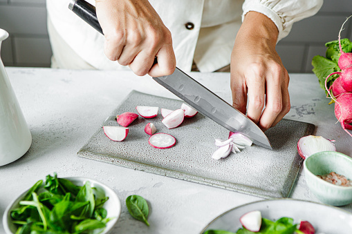 Cropped shot of hands cut radishes with a kitchen knife on a cutting board. Close-up of woman chopping radishes over kitchen counter.
