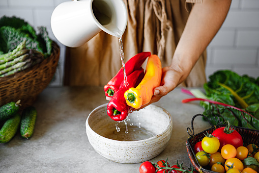 Close-up of woman hands washing red and yellow peppers over a bowl with mug of water. Female washing vegetables over kitchen counter.