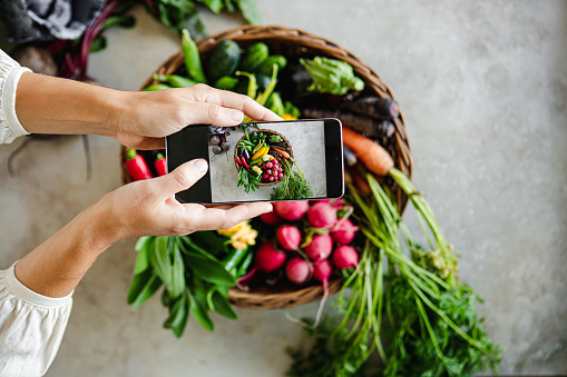 Point of view of a woman hand holding smartphone and taking photos of basket full of fresh vegetables on table. Close-up of woman photographing variety fresh and healthy vegetable with her mobile phone on kitchen table.