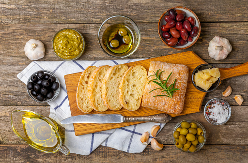 Ciabattas, olive oil in a bowl with olives, herbs, spices, garlic, pesto, parmesan and ciabatta bread on a texture background.Banner. Healthy food concept.Delicacy. Mediterranean Kitchen. Copy space.