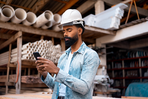 Civil engineer with mobile phone. Confident Professional black man with helmet texting and typing on the mobile smart phone in construction warehouse