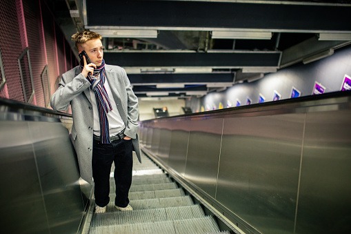 Teenage boy on his way up standing in the subway on escalator in Stockholm, Sweden. He using mobile phone and wears elegant coat and scarf in early spring.