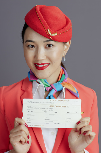 happy modern asian female air hostess in red skirt, jacket and hat uniform with flight tickets isolated on gray.