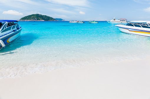 A tropical beach of Similan islands with white sand, clear water and boats. The water is a beautiful turquoise color and is very clear. There are several boats in the water, some are small and blue and white, while others are larger and white. The boats are anchored near the shore. In the background, there is a small island with green trees and a rocky coastline. The sky is blue and clear. This photo is a perfect example of a tropical paradise.