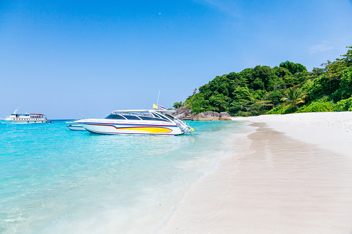 A tropical beach of similan islands with trees, rocks and boats in clear water. The beach has white sand and is lined with trees and rocks. There are two boats in the water, one is a white and blue speedboat and the other is a larger white boat with a blue stripe. The water is a bright blue and is very clear. The sky is a bright blue with a few clouds. This photo creates a sense of peace and relaxation.