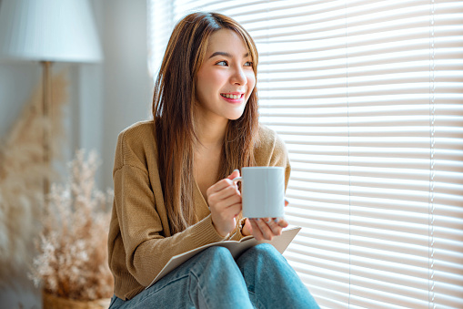 Joyful young asian female enjoying a cup of coffee while sitting on bench at home, Cosy scene, Smiling pretty woman drinking hot tea in autumn winter.