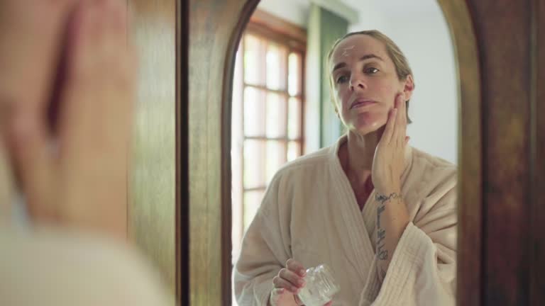 Woman in a bathrobe applying cream to her face in a bedroom mirror