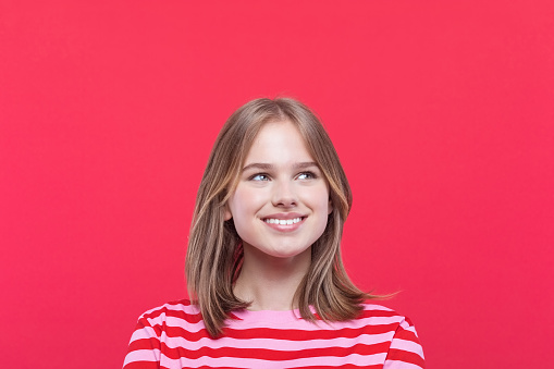 Happy teenage girl wearing striped pink t-shirt looking away and smiling. Studio shot, red background.