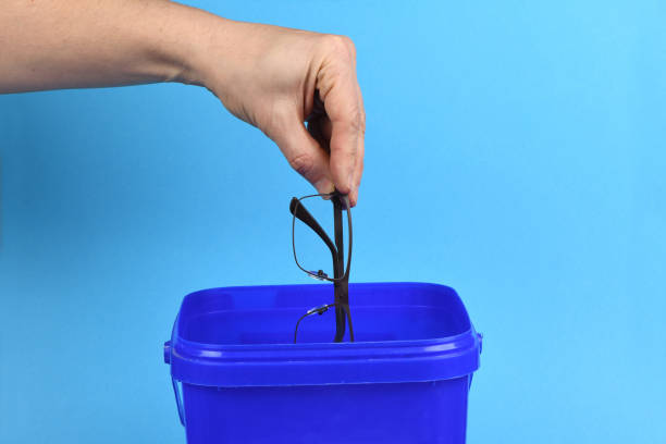 istock Hand holding plastic black glasses puts in blue container, isolated on white background. 1644709642