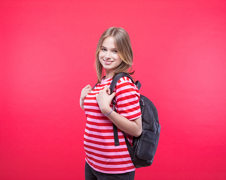 Happy female high school student wearing striped pink t-shirt and black backpack smiling at camera. Studio shot, red background.