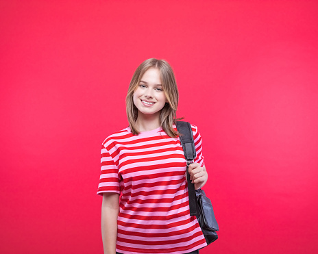 Happy female high school student wearing striped pink t-shirt and black backpack smiling at camera. Studio shot, red background.