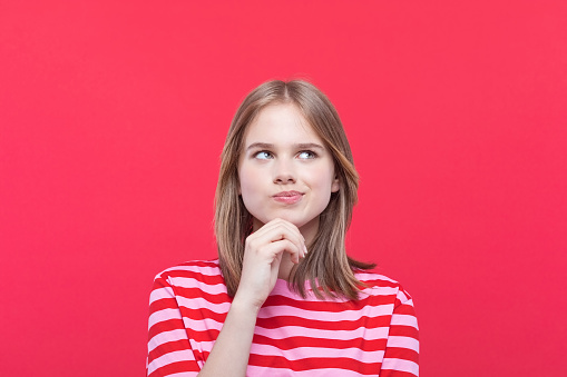 Thoughtful teenage girl wearing striped pink t-shirt looking away with hand on chin. Studio shot, red background.