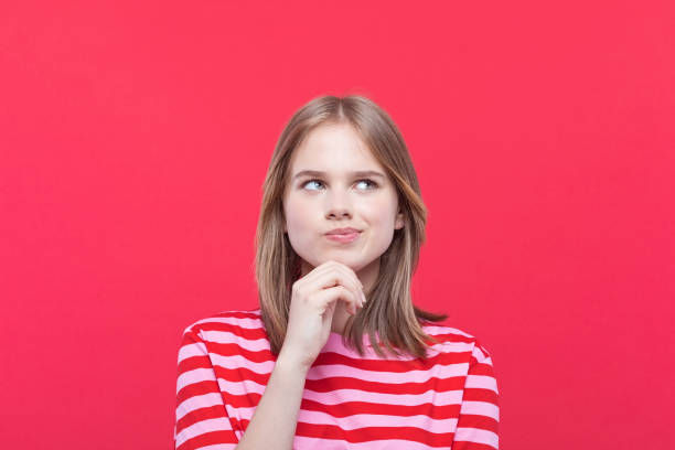 Portrait of confused teenage girl Thoughtful teenage girl wearing striped pink t-shirt looking away with hand on chin. Studio shot, red background. skeptical look stock pictures, royalty-free photos & images