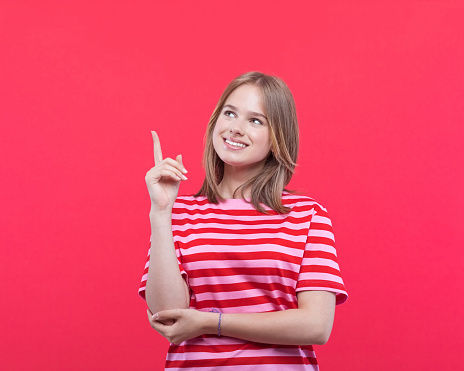 Happy teenage girl wearing striped pink t-shirt pointing at copy space with index finger. Studio shot, red background.