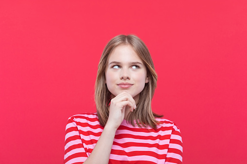 Pensive teenage girl wearing striped pink t-shirt looking away with hand on chin. Studio shot, red background.