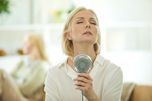 Menopausal mature woman sitting on sofa in the living room at home having a hot flash and using fan. Her displeased teenage girl sitting in the background. They are in conflict.
